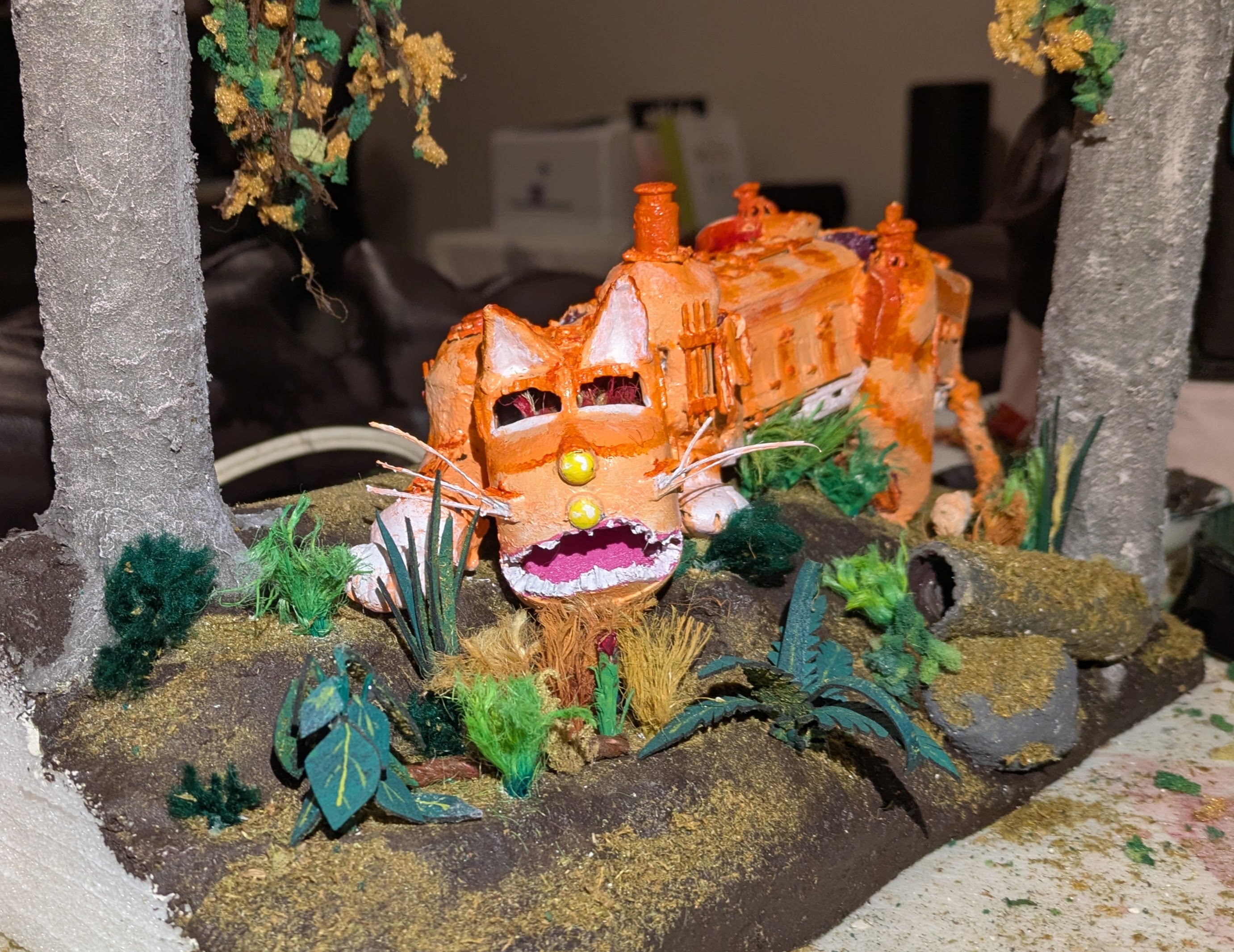 A toy train modified with cat ears, paws, and tail. It is painted like an orange tabby cat, it is surrounded by low foliage and framed by two grey tree trunks, it is posed as though it's stalking prey.